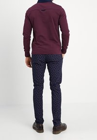 Bordeaux langarmshirt met een marineblauwe kraag, gecombineerd met goed passende marineblauwe broek met een rood cirkelpatroon. Bruine schoenen maken de look compleet.