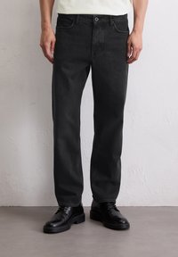 Mężczyzna w czarnych dżinsach o prostym kroju i czarnych skórzanych butach na sznurowadłach stoi na szarym podłodze przy białej ścianie.