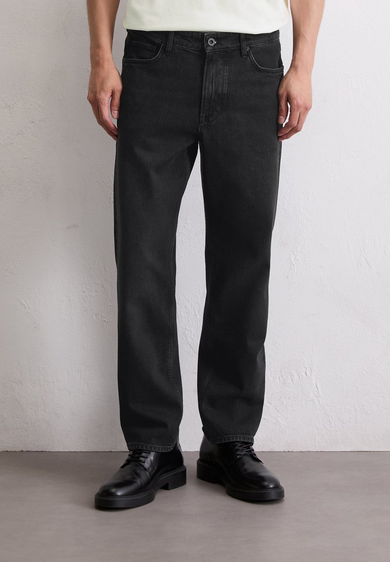 Mężczyzna w czarnych dżinsach o prostym kroju i czarnych skórzanych butach na sznurowadłach stoi na szarym podłodze przy białej ścianie.