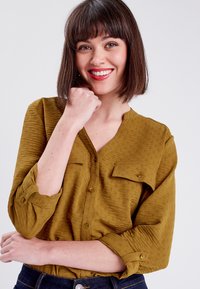Chemise boutonnée couleur moutarde avec un tissu texturé, dotée de deux poches poitrine et de manches avec pattes de roulage. Encolure simple et patte de boutonnage avant.