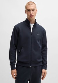 Marinblå zip-up sweatshirt med hög krage, sidofickor, slät textur och en diskret logotyp på bröstet. Matchad med byxor i samma färg.