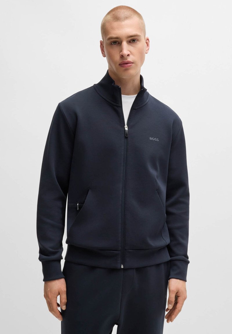 Marinblå zip-up sweatshirt med hög krage, sidofickor, slät textur och en diskret logotyp på bröstet. Matchad med byxor i samma färg.