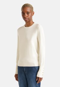 Crewneck-Pullover in Creme, aus weichem Stoff gefertigt. Verfügt über lange Ärmel mit goldfarbenen Knopf-Details an den Bündchen. Kombiniert mit blauen Jeans.