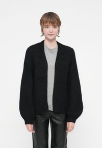 Cardigan nero lavorato a maglia con una vestibilità rilassata, apertura frontale e maniche voluminose. Indossato sopra una maglia grigia, abbinato a pantaloni neri lucidi.