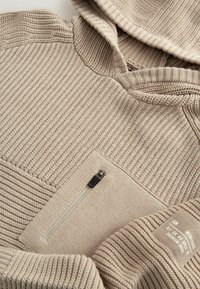 Hoodie beige avec un motif tricoté texturé, une poche poitrine zippée et des poignets côtelés. Les accents comprennent une étiquette logo sur la manche.