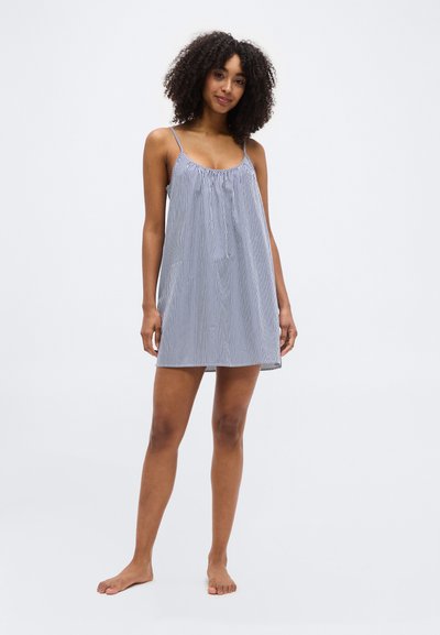 Γυναίκα με κατσαρά μαλλιά που στέκεται ξυπόλητη, φορώντας ένα φαρδύ, αμάνικο κοντό φόρεμα με μπλε και λευκές ρίγες, μπροστά σε λευκό φόντο.