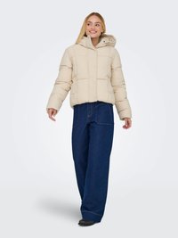 Casaco puffer bege com fecho de botão e capuz, apresentando um design acolchoado. Combinado com calças de ganga azul-escuro de corte largo.