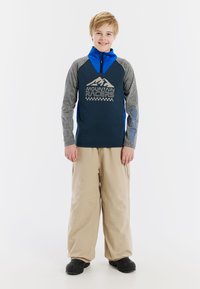 Camiseta de manga larga azul y gris con gráfico de "Mountain Racers", combinada con pantalones de nieve beige y botas negras, de pie sobre un fondo blanco.