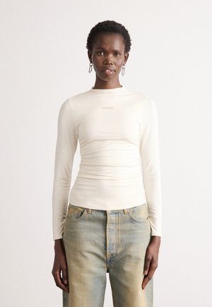 Kvinne i en tettsittende kremfarget langermet topp med "DKNY"-logo og lyseblå høyhælte jeans, stående foran en ensfarget hvit bakgrunn.