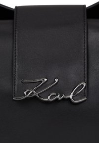 Zwarte leren tas met klep, voorzien van een zilverkleurig metalen scriptlogo met de tekst "Karl" geborduurd op de voorkant.