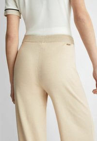 Pantaloni beige a gamba larga con una leggera lucentezza, texture morbida e una fascia in vita accentuata da dettagli dorati. Vista posteriore che mostra linee fluide e cuciture dettagliate.