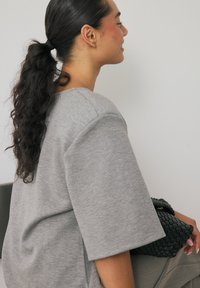 Pilkas, didelis sportinis megztinis su trumpomis rankovėmis, tekstūruota medžiaga ir laisvas kirpimas. Juoda pintinė rankinė laikoma rankoje. Neutralus fonas.