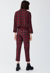 Salopette en tissu à carreaux rouges et noirs avec une coupe décontractée, des manches longues, une taille élastique et des jambes fuselées. Portée avec des bottines noires.