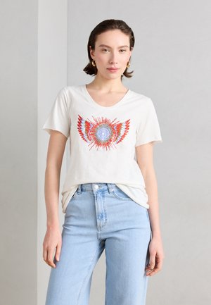 Nuori nainen, jolla on lyhyet ruskeat hiukset, kullanväriset renkaankorvakorut, valkoinen T-paita, jossa on värikäs auringonsäteiden kuvio, ja vaaleansiniset farkut.