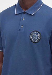 Polo bleu avec trois boutons et un écusson du logo Milan sur le côté gauche de la poitrine, porté par une personne à la peau foncée.