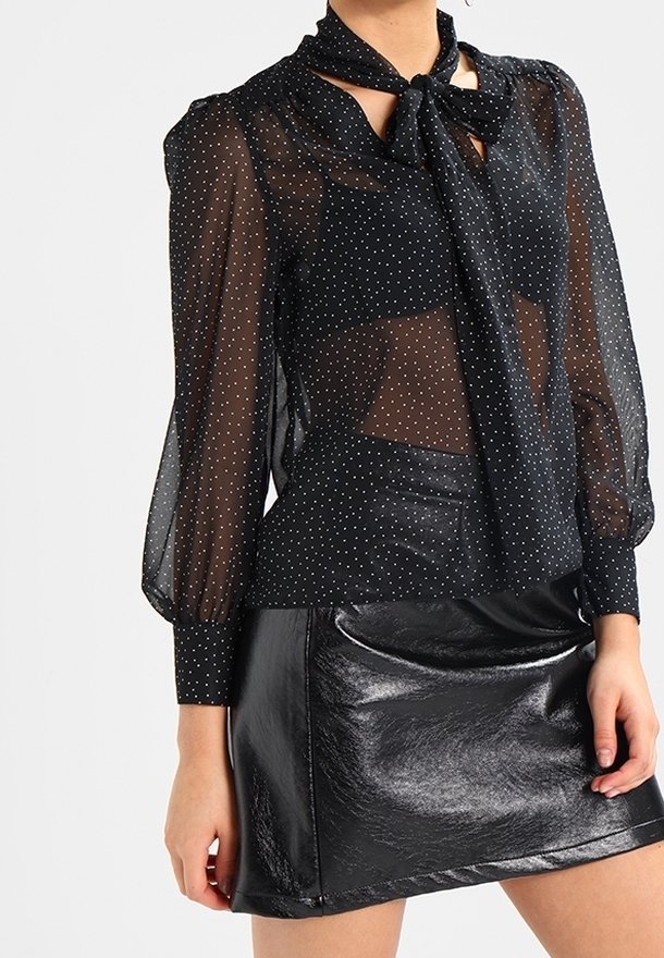 Blouse noire transparente à pois blancs avec une cravate, associée à une mini-jupe en cuir noire brillante sur une personne debout.