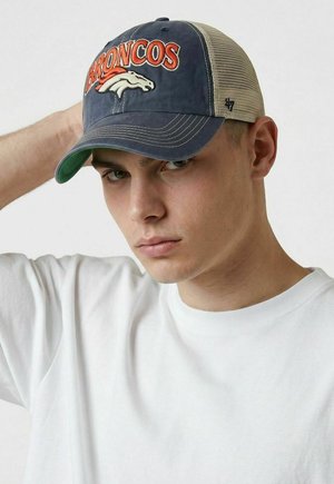 Young man wearing a Denver Broncos cap with navy front, beige mesh back, and white T-shirt, looking thoughtfully at the camera.