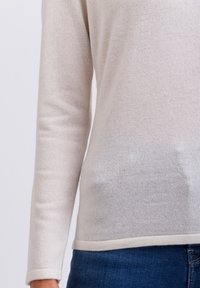 Helle beigefarbene Pullover mit langen Ärmeln, weicher Strickstruktur und Rundhalsausschnitt. Am unteren Rand mit einem kontrastierenden Bund akzentuiert. Über blauen Jeans getragen.