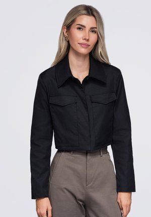 Femme aux longs cheveux blonds, portant une veste noire courte et un pantalon taille haute couleur taupe, debout devant un fond clair uni.