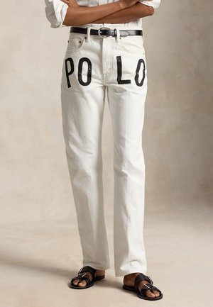 Άτομο που φοράει άσπρο τζιν με μεγάλο μαύρο κείμενο "PO LO" μπροστά, μαύρη ζώνη, λευκό πουκάμισο με γυρισμένα μανίκια και μαύρα σανδάλια.