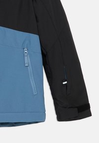Kleurgeblokte jas met een zwart bovenstuk en een blauw onderstuk, voorzien van een ritszak en klittenbandmanchetten. Gladde textuur.