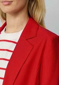 Rode blazer met een gestructureerde revers, een gladde textuur en een getailleerd ontwerp, gecombineerd met een wit shirt met rode strepen.