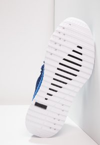 Niebieska podeszwa buta sportowego z fakturowaną białą powierzchnią, wyposażona w rowki i czarny akcent, zaprojektowana dla przyczepności i elastyczności.