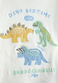 Żółty stegozaur, zielony T-Rex i niebieski triceratops wyhaftowane wokół napisów "Dino Bedtime Club" i "Snoozesaurus" na białej tkaninie.