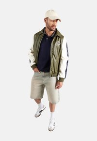 Chaqueta bomber de cuero verde oliva con mangas crema y azul marino, combinada con una polo azul marino, pantalones cortos de mezclilla claros y zapatillas blancas.