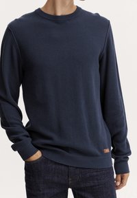 Pull en tricot bleu marine avec un motif texturé, col rond et poignets côtelés, complété par un petit patch logo en cuir en bas à gauche.