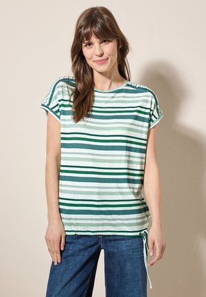 Camiseta de manga corta a rayas en tonos verdes y blancos, con detalles fruncidos en los hombros y un dobladillo con cordón. Combinada con pantalones de mezclilla.