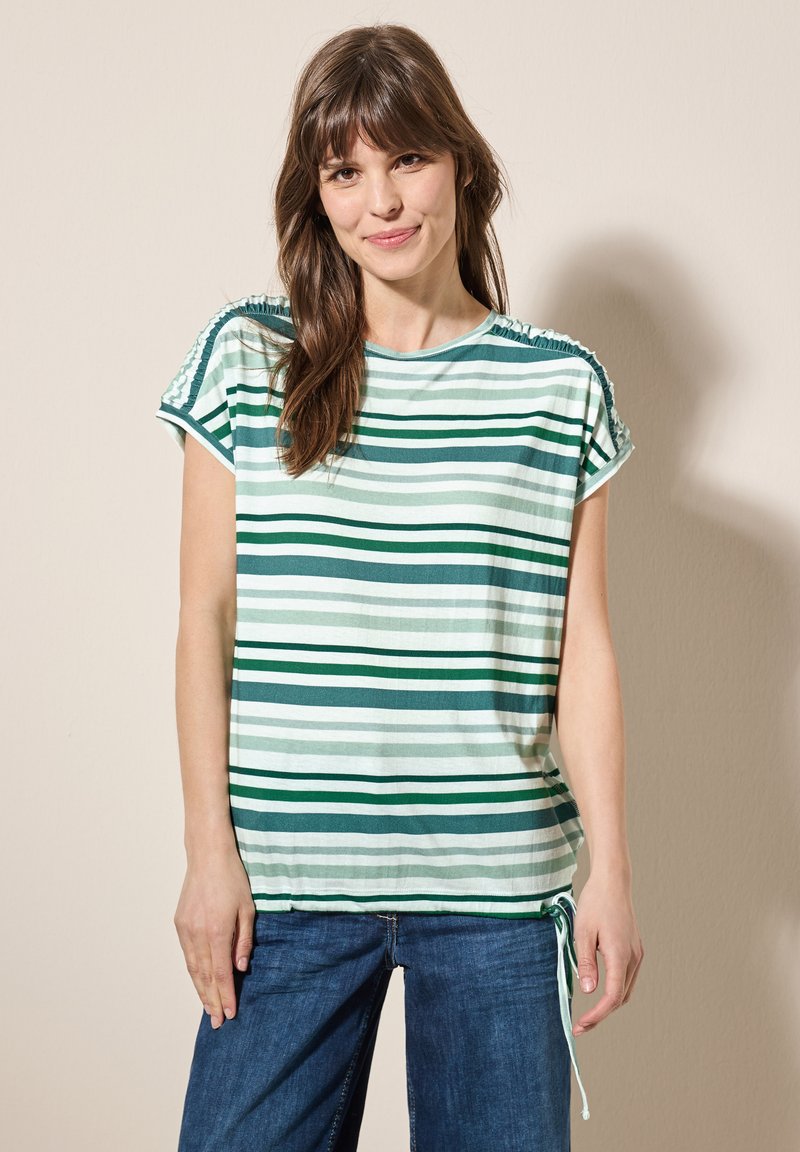 Camiseta de manga corta a rayas en tonos verdes y blancos, con detalles fruncidos en los hombros y un dobladillo con cordón. Combinada con pantalones de mezclilla.