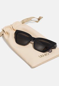 Occhiali da sole neri angolari con una robusta montatura in plastica appoggiati su una sacca in tessuto beige con coulisse e il marchio "LIU-JO".