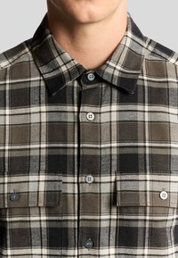 Mand iført en knaplukket brun, sort og beige plaid flannel skjorte med krave og brystlommer foran en ensfarvet baggrund.