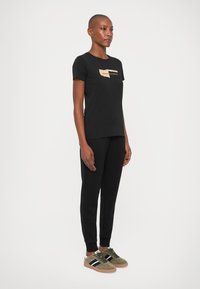 Tricou negru cu un logo bej, asortat cu pantaloni de trening negri și adidași verde măsliniu cu dungi bleumarin. Design casual, ajustat.