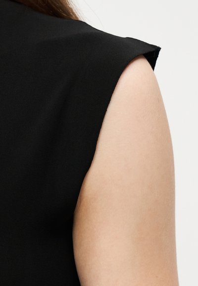 Αμάνικη μαύρη μπλούζα που δείχνει το πάνω μέρος του μπράτσου και τον ώμο πάνω σε απλό ανοιχτό φόντο, με ορατή υφή στο ύφασμα.