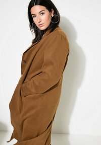 Bruine wolmixjas met een geperforeerde revers, twee voorzakken en een enkelzijdige knoopsluiting; zachte textuur en oversized pasvorm.