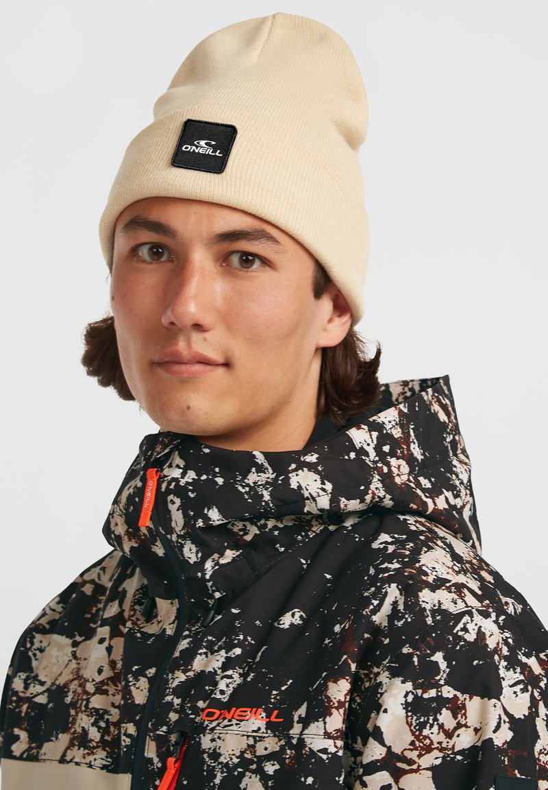 Beige hues beanie med et sort brandmærke, parret med en sort og hvid jakke med sprøjtet mønster og orange lynlåse.