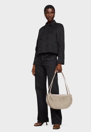 Frau trägt eine schwarze, kurz geschnittene, doppelreihige Jacke, schwarze Jeans mit weitem Bein, schwarze Sandalen und hält eine beigefarbene Wildleder-Handtasche mit zwei Trägern.