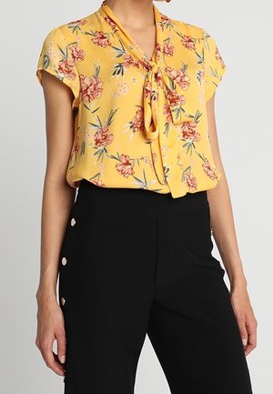 Frau trägt eine gelbe, kurzärmelige Bluse mit Blumenmuster und Halsband, kombiniert mit hoch taillierter schwarzer Hose mit seitlichen Knöpfen.