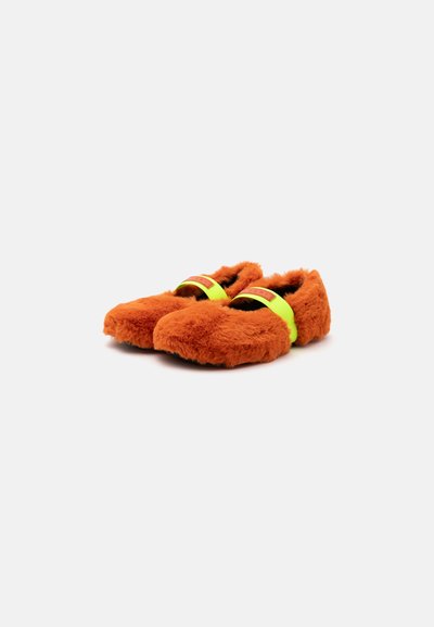 Paire de chaussons orange duveteux avec une sangle jaune vif sur le dessus, posés sur un fond clair uni.