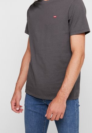 Mann trägt ein dunkelgraues Levi's T-Shirt mit rotem Logo auf der Brust und blaue Jeans, steht vor einem schlichten hellen Hintergrund.