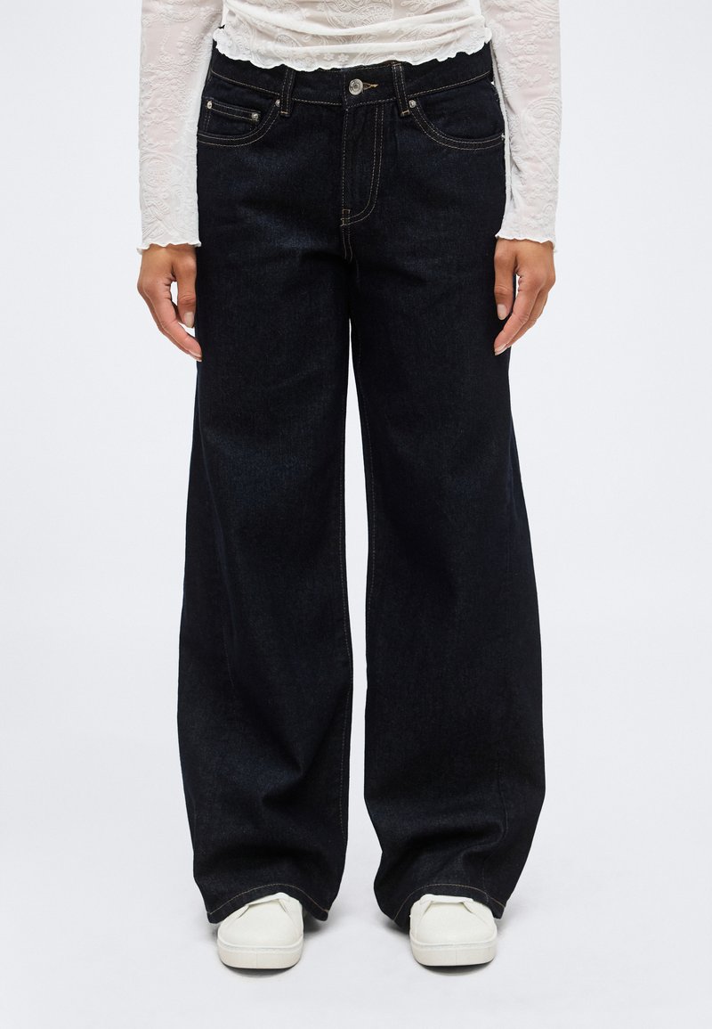 Άτομο που φοράει μαύρο παντελόνι με φαρδιά μπατζάκια, λευκό μακρυμάνικο τοπ με υφή και λευκά αθλητικά παπούτσια, στέκεται μπροστά σε απλό φόντο.