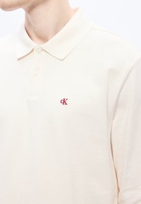 Gaiši bēša polo krekls no teksturētas auduma. Tam ir trīs pogu plāksne un neliels sarkans "CK" logotips kreisajā krūtīs.