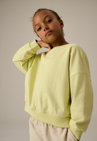 Jasnożółty, oversize'owy sweatshirt z ściągaczami przy rękawach i u dołu. Miękka faktura, okrągły dekolt, w zestawieniu z jasnobeżowymi spodniami.