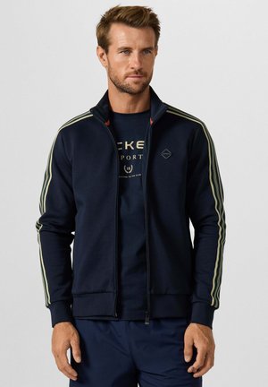 Man draagt een donkerblauw jack met rits met beige strepen op de mouwen en bijpassende donkerblauwe broek, staand tegen een witte achtergrond.