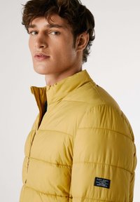 Chaqueta acolchada de color amarillo claro con cuello alzado y costuras horizontales. Cuenta con un cierre lateral y un parche de marca negro en la manga.