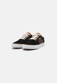 Black suede sneakers with cream laces and a brown accent panel, featuring a white rubber sole and an orange logo on its tongue.