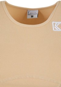 Camiseta sin mangas de color beige con un escote redondo, que presenta una etiqueta tejida marcada con "S" y un pequeño acento de logo blanco cerca del hombro.