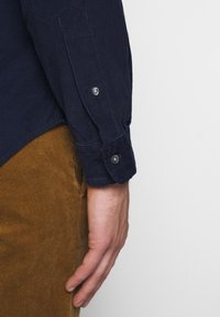 Námořnická kordurová košile s knoflíkovými manžetami, vyrobená z texturované látky. Hnědé kordurové kalhoty viditelné ve spodní části obrázku.